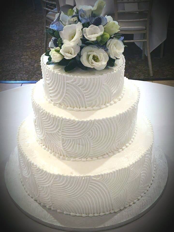 Three-tier white cake with patterned icing and a bouquet of white and blue flowers
